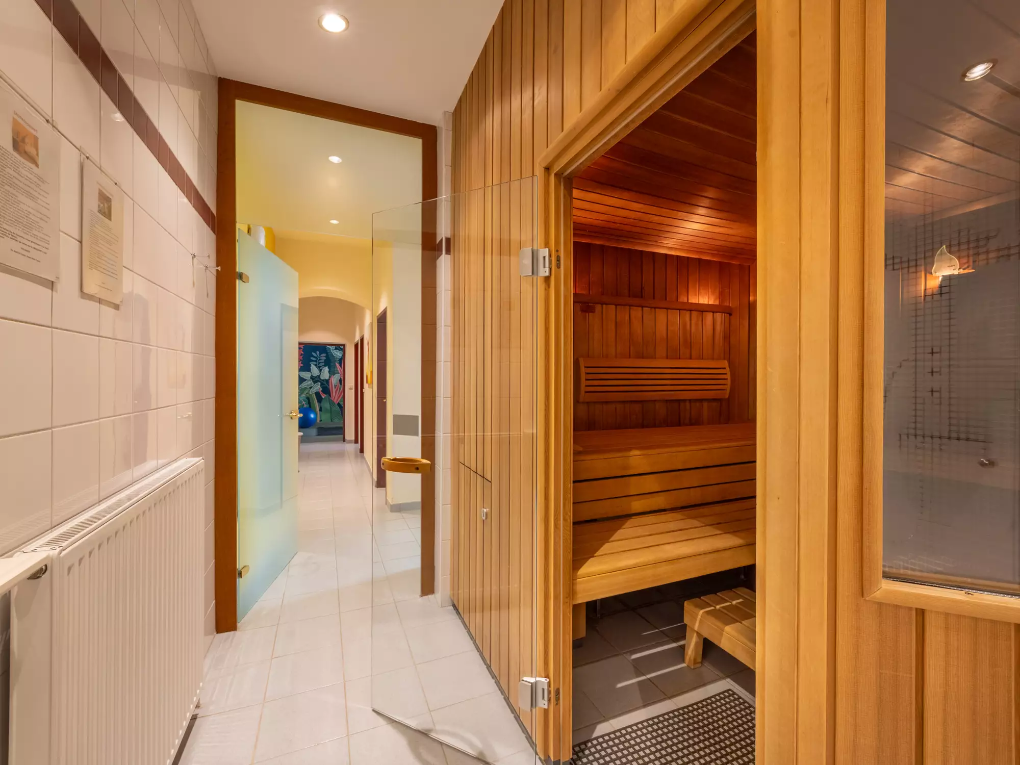 Ein heller Flur mit einer Glastür führt zu einer Sauna aus hellem Holz. Im Hintergrund ist ein Badezimmer mit einer bunten Wanddekoration sowie weiteren Türen zu sehen. An der Wand hängen Informationsblätter.
