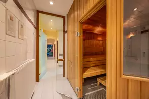 Ein heller Flur mit einer Glastür führt zu einer Sauna aus hellem Holz. Im Hintergrund ist ein Badezimmer mit einer bunten Wanddekoration sowie weiteren Türen zu sehen. An der Wand hängen Informationsblätter.