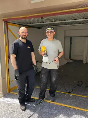 Zwei Männer stehen in einem Raum, der teilweise renoviert wird. Der Mann links trägt ein schwarzes T-Shirt und hat einen Bart, der rechte Mann trägt ein graues Shirt und hält ein Sprühgerät. Beide tragen Handschuhe und lächeln in die Kamera.