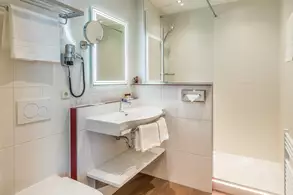 Ein modernes Badezimmer mit einem weißen Waschbecken, einem großen Spiegel, einem Handtuchhalter und einer Dusche. Die Wände sind hell gefliest, und es gibt eine Handtuchablage sowie einen Toilettenbereich. Die Beleuchtung ist angenehm und sorgt für eine helle Atmosphäre.