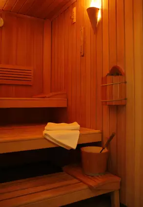 Innenansicht einer Sauna mit Holzwänden und einer Bank. Auf der oberen Bank liegt ein gefaltetes Handtuch und daneben steht ein Eimer mit einem Schöpflöffel. Eine Wandlampe sorgt für sanftes Licht.