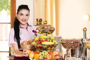 Eine Frau mit dunklen Haaren und rotem Lippenstift steht an einem Buffet und hält eine Schale mit Obst. Neben ihr befinden sich weitere Obstschalen und Nüsse. Sie trägt ein gestreiftes Hemd und lächelt freundlich in die Kamera.