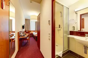 Blick in ein Hotelzimmer, das einen Eingangsbereich zeigt. Auf der linken Seite ein Spiegel, rechts eine Dusche und ein Waschbecken. Im Hintergrund sind ein Tisch, Stühle und ein Bett sichtbar. Tageslicht strömt durch die Fenster, die mit Vorhängen dekoriert sind.