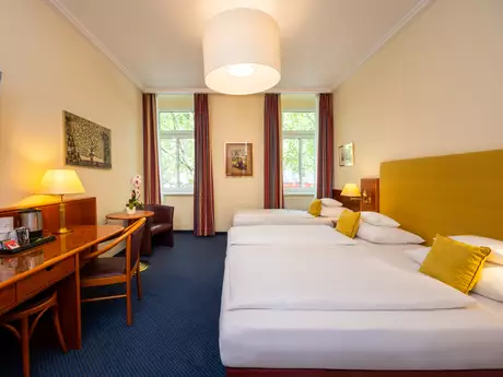 Ein Hotelzimmer mit einem großen Doppelbett und einem Einzelbett und gelben Kissen. Es gibt einen Tisch mit Stühlen, eine Lampe und einen Fernseher an der Wand. Fenster mit Vorhängen lassen Tageslicht hereinfällen. An der Wand hängen Bilder und eine Orchidee steht auf dem Tisch.