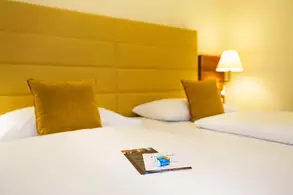 Zwei Betten in einem Hotelzimmer mit gelben Kopfenden und Kissen. Auf einem Bett liegt eine Broschüre oder ein Flyer. An der Wand befindet sich eine Lampe, die warmes Licht spendet.