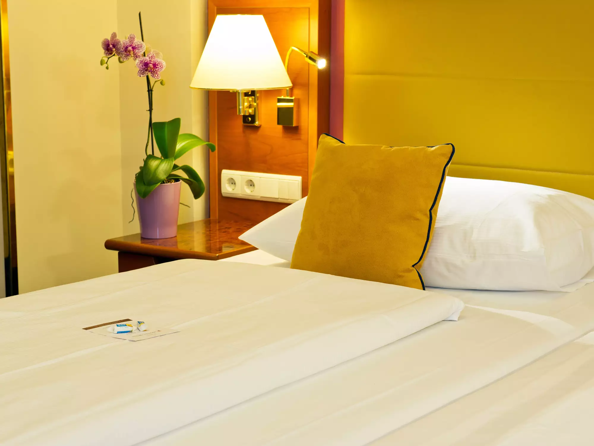 Das Bild zeigt ein Schlafzimmer mit einem Bett, auf dem ein gelbes Kissen liegt. Neben dem Bett steht ein Holztisch mit einer Pflanze und einer Lampe. Im Hintergrund sind gelbe Wände zu sehen.