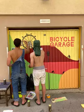 Zwei Männer, die mit Farben eine Garagentür gestalten. Die Tür hat das Motiv "Bicycle Garage" und zeigt bunte Landschaften sowie Fahrräder. Einer der Männer trägt einen Overall, der andere Shorts. Beide sind oberkörperfrei und arbeiten konzentriert an der Wandgestaltung.