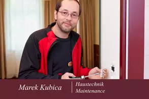 Ein Mann mit Brille und leichtem Bart steht in einem Raum und hält einen Schraubenschlüssel in der Hand. Er trägt ein rotes und schwarzes Oberteil und lächelt. Im Hintergrund sind Vorhänge und eine Wand zu sehen.