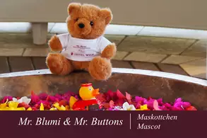 Ein brauner Teddybär namens Mr. Blumi sitzt neben einer kleinen gelben Gummiente namens Mr. Buttons in einer Schüssel mit bunten Blütenblättern. Der Teddybär trägt ein weißes T-Shirt mit der Aufschrift „Hotel Wien“.