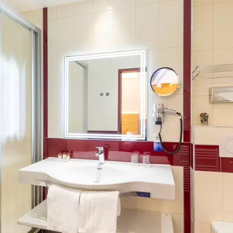 Das Bild zeigt ein modernes Badezimmer mit einer Dusche, einem Waschbecken und einem großen Spiegel. Die Wände sind hell gefliest, und es gibt rote Akzente. Handtücher liegen auf einem Regal.