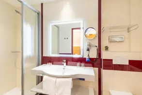 Das Bild zeigt ein modernes Badezimmer mit einer Dusche, einem Waschbecken und einem großen Spiegel. Die Wände sind hell gefliest, und es gibt rote Akzente. Handtücher liegen auf einem Regal.