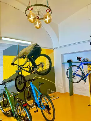 In einem modernen Raum sind mehrere Fahrräder aufgestellt. An der Wand ist ein BMX-Fahrer abgebildet, der einen Trick ausführt. Die Wände sind in verschiedenen Farben gestrichen, und eine kreative Lampeninstallation hängt von der Decke. Der Boden ist orange.