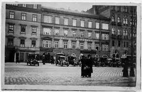 Ein historisches Stadtszenario mit dem Hotel Nordbahn im Hintergrund. Vor dem Gebäude stehen mehrere Autos, während zwei Personen in schwarzer Kleidung im Vordergrund stehen. Die Straße ist mit Kopfsteinpflaster bedeckt.