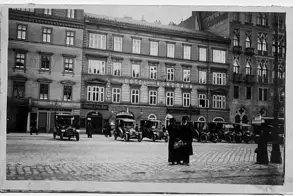 Ein historisches Stadtszenario mit dem Hotel Nordbahn im Hintergrund. Vor dem Gebäude stehen mehrere Autos, während zwei Personen in schwarzer Kleidung im Vordergrund stehen. Die Straße ist mit Kopfsteinpflaster bedeckt.