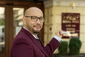 Ein Mann in einem burgunderfarbenen Anzug steht vor einem Gebäude und zeigt mit dem Finger auf ein Schild. Das Schild trägt den Schriftzug des Hotels. Im Hintergrund sind große Fenster und einige Pflanzen sichtbar.