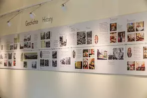 Eine Wand mit zwei Sprachen beschriftet: "Geschichte" und "History". Daneben eine Vielzahl von Bildern und Texten, die historische Ereignisse und Informationen darstellen, gegliedert nach Jahren.