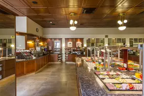 Ein Buffet-Restaurant mit einem Holzdekor. Es gibt mehrere Buffets mit einer Auswahl an belegten Broten, frischem Obst und Gemüse. Die Atmosphäre ist hell und einladend, mit Tischlampen und einer eleganten Deckenverkleidung.
