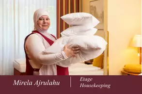Eine Frau in einem roten Oberteil und mit einem Kopftuch hält mehrere Kissen in ihren Armen. Sie steht in einem hellen Raum mit einem Bett im Hintergrund. Der Text am unteren Rand nennt ihren Namen und die Abteilung „Housekeeping“.