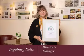 Frau Ingeborg Seitz steht lächelnd in einem Raum mit historischen Bildern an der Wand. Sie hält ein gerahmtes Zertifikat in der Hand. Im Hintergrund sind gemütliche Sitzgelegenheiten und eine Pflanze zu sehen.