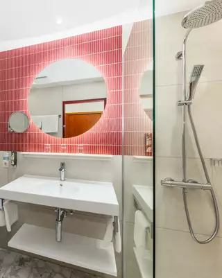 Ein modernes Badezimmer mit einem Waschbecken, einem großen Spiegel und einer Duschkabine. Die Wände sind in hellen Farbtönen gehalten, und eine hervorgehobene Wand hat pinke Fliesen. Es gibt Handtücher, Toilettenpapier und ein Handtuchhalter aus Edelstahl.