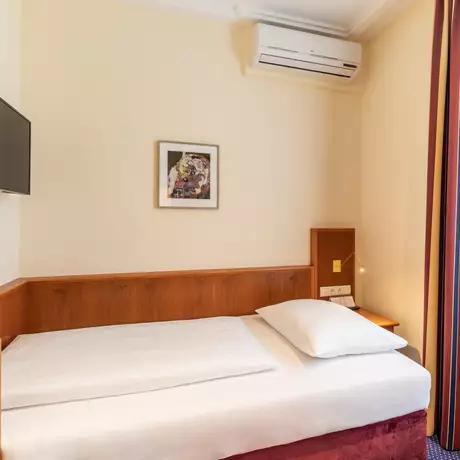 Ein einfaches Hotelzimmer mit einem Einzelbett, einem kleinen Fernseher an der Wand, einem Tisch und einer Lampe. An den Fenstern hängen Vorhänge mit Streifen. Die Wände sind hell gefärbt und es gibt eine Klimaanlage.