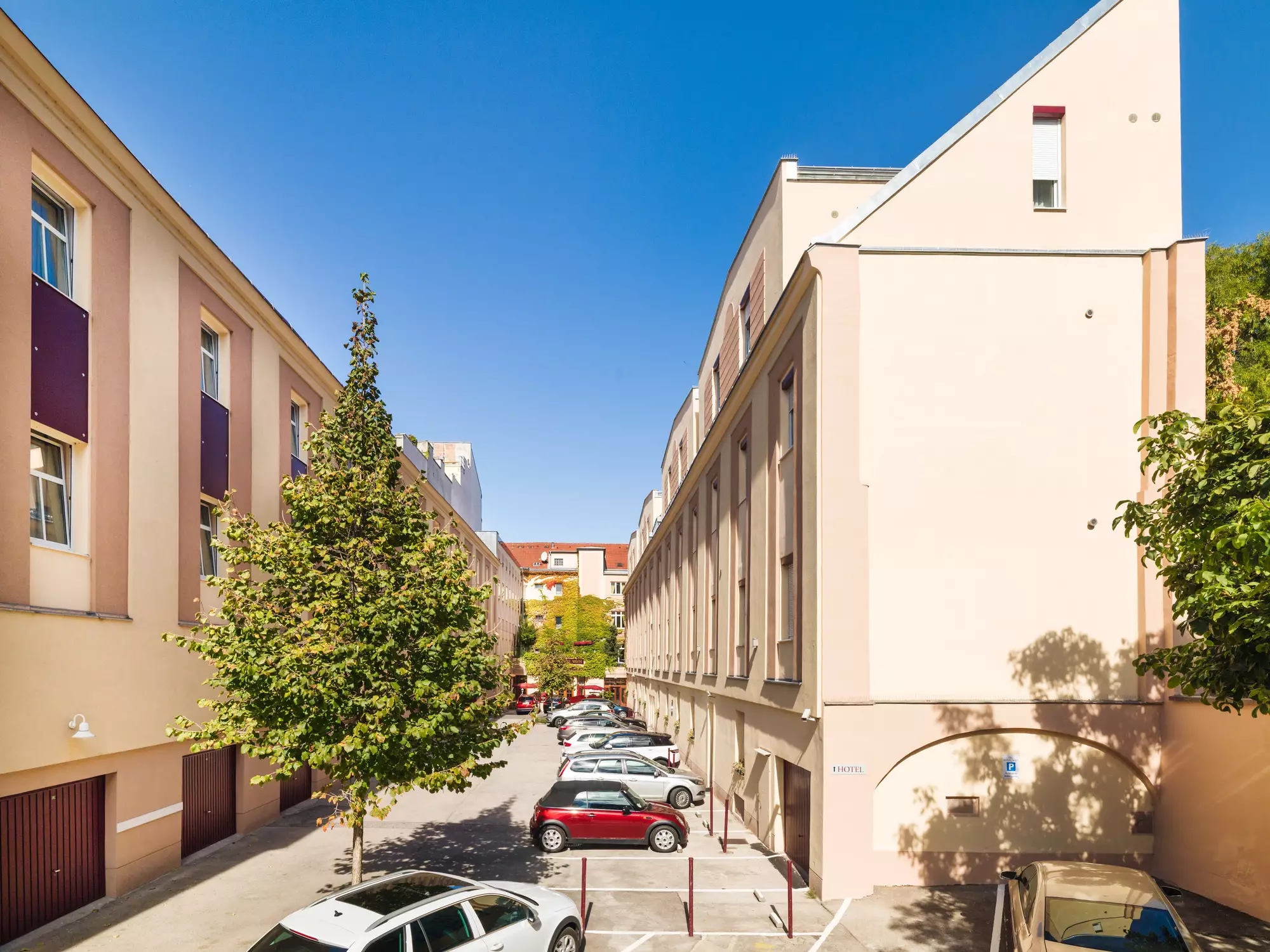 Eine helle Stadtszene zeigt eine Parkanlage zwischen Wohngebäuden. Mehrere Autos stehen auf einem Parkplatz, umgeben von Grünanlagen und Bäumen. Der Himmel ist klar und blau, was eine freundliche Atmosphäre vermittelt.