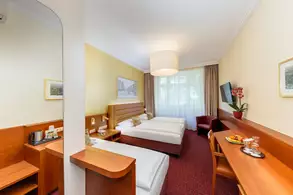 Ein gemütliches Hotelzimmer mit zwei Einzelbetten, einem Schreibtisch und einem Stuhl. An der Wand hängen Bilder, und ein großer Fenster mit Vorhängen lässt Tageslicht herein. Auf dem Tisch sind Snacks und Getränke platziert.