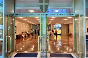 Der Eingang zum historischen Museum der Stadt Wien ist sichtbar, mit großen Glastüren, die offenstehen. Im Inneren sind Besucher zu sehen, die sich im Foyer bewegen. Der Boden ist glänzend und reflektiert das Licht.