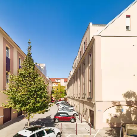 Blick auf einen ruhigen Innenhof mit parkenden Autos, umgeben von modernen Gebäuden. Auf der linken Seite steht ein Baum, während der Himmel klar und blau ist. Die Umgebung wirkt sauber und gut beleuchtet.