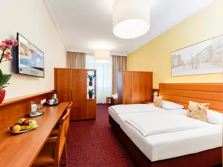 Ein gemütlich eingerichtetes Hotelzimmer mit zwei Einzelbetten, einer Holzkommode, einem Tisch mit Obstkorb und einem großen Fenster. An der Wand hängt ein Fernseher und es gibt eine Pflanze auf dem Tisch. Der Boden ist mit rotem Teppich ausgelegt.