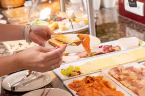 Eine Hand hält einen Teller mit zwei Brotscheiben und einem Stück geräuchertem Lachs. Im Hintergrund ist ein Buffet mit verschiedenen Käsesorten, Aufschnitt und Obst sichtbar.