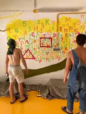 Zwei Personen stehen vor einer bunten Wand, die mit Zahlen und Buchstaben in verschiedenen Farben bemalt ist. Eine Person trägt eine grüne Kopfbedeckung und hat eine Spraydose in der Hand. Die andere Person trägt einen Latzhose. Im Vordergrund liegen Abdeckplanen.