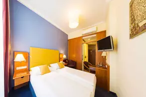 Ein modern eingerichtetes Hotelzimmer mit zwei Betten, einem gelben Kopfteil und dekorativen Kissen. An der Wand befinden sich ein Fernseher und eine weitere Möbelstück mit einer Lampe. Die Wände sind grau und cremefarben, mit einem schönen Licht von der Decke.