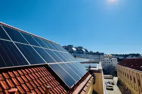 Auf einem Dach sind Solarpaneele montiert, die Sonnenlicht einfangen. Im Hintergrund erstreckt sich eine städtische Landschaft unter einem klaren blauen Himmel mit der Sonne strahlend am oberen Rand des Bildes. Die Umgebung ist ruhig mit einigen parkenden Autos.