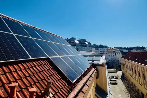 Auf einem Dach sind Solarpaneele montiert, die Sonnenlicht einfangen. Im Hintergrund erstreckt sich eine städtische Landschaft unter einem klaren blauen Himmel mit der Sonne strahlend am oberen Rand des Bildes. Die Umgebung ist ruhig mit einigen parkenden Autos.