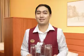 Ein Mitarbeiter in einem Hotelzimmer trägt eine Karaffe mit Wasser und mehrere Gläser. Er trägt eine rote Weste über einem weißen Hemd. Der Hintergrund zeigt eine orangefarbene Wand und eine verschwommene Fotografie einer Stadtansicht.