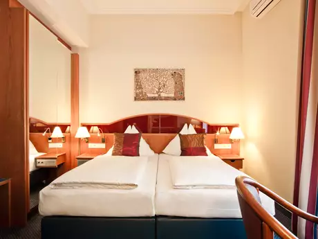 Das Bild zeigt ein modernes Hotelzimmer mit zwei Einzelbetten, einer gemütlichen Einrichtung und einem Spiegel. An der Wand hängt ein Kunstwerk. Auf den Nachttischen stehen Lampen, und das Zimmer wirkt hell und einladend.