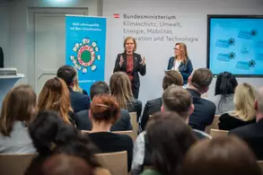 Eine Veranstaltung im Bundesministerium für Klimaschutz, Umwelt, Energie, Mobilität, Innovation und Technologie. Eine Rednerin steht im Vordergrund und spricht, während eine weitere Person neben ihr zuhört. Im Hintergrund sind Plakate und Präsentationen sichtbar. Das Publikum sitzt und hört aufmerksam zu.