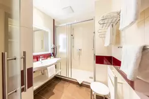 Ein modernes Badezimmer mit einer Dusche, einem Waschbecken und einem großen Spiegel. Weiße Handtücher sind auf einem Regal und an einem Handtuchhalter aufgehängt. Die Wände sind hell gefliest, mit roten Akzenten. Der Boden ist dunkelbraun.