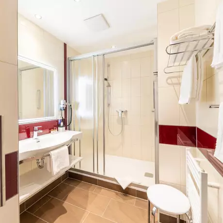 Ein modernes Badezimmer mit einer Dusche, einem Waschbecken und einem großen Spiegel. Weiße Handtücher sind auf einem Regal und an einem Handtuchhalter aufgehängt. Die Wände sind hell gefliest, mit roten Akzenten. Der Boden ist dunkelbraun.