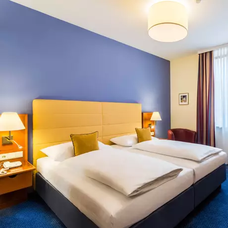 Ein helles Hotelzimmer mit zwei Einzelbetten, einem Tisch mit Stuhl, einer großen Fensterfront mit Vorhängen und einem Flachbildfernseher an der Wand. Nachttische mit Lampen stehen neben den Betten. Die Wände sind in sanften Farben gestaltet.