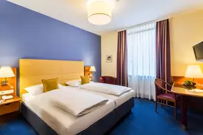 Ein helles Hotelzimmer mit zwei Einzelbetten, einem Tisch mit Stuhl, einer großen Fensterfront mit Vorhängen und einem Flachbildfernseher an der Wand. Nachttische mit Lampen stehen neben den Betten. Die Wände sind in sanften Farben gestaltet.
