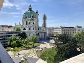 Blick auf einen Platz in Wien mit der imposanten Karlskirche im Hintergrund. Umgeben von Bäumen und Grünflächen, sind Menschen auf dem Platz zu sehen. Das Wetter ist sonnig und der Himmel klar. Ein Gebäude im Hintergrund wird renoviert.