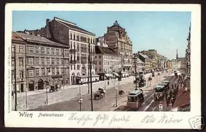 Straßenansicht der Proleterstraße in Wien, mit historischen Gebäuden, Straßenbahnen und Fahrzeugen. Die Szene vermittelt einen Eindruck des urbanen Lebens und der Architektur der Zeit. Es herrscht lebhaftes Treiben auf der Straße.