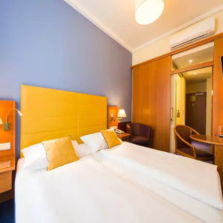 Ein Hotelzimmer mit zwei Einzelbetten und gelben Kissen. An der Wand ist ein großer, gelber Kopfteil zu sehen. Es gibt einen Tisch mit einem Stuhl, eine Lampe und einen Fernseher. Im Hintergrund ist eine Tür zu einem Badezimmer oder einem anderen Raum sichtbar.
