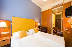 Ein Hotelzimmer mit zwei Einzelbetten und gelben Kissen. An der Wand ist ein großer, gelber Kopfteil zu sehen. Es gibt einen Tisch mit einem Stuhl, eine Lampe und einen Fernseher. Im Hintergrund ist eine Tür zu einem Badezimmer oder einem anderen Raum sichtbar.