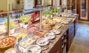 Reichhaltiges Frühstücksbuffet mit frischem Gebäck, Obstplatten und warmen Speisen in hellen Räumlichkeiten.