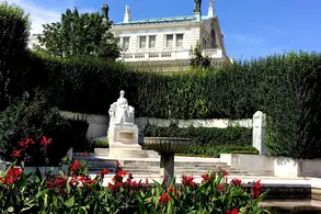 Ein Garten mit blühenden roten Blumen im Vordergrund, einem Springbrunnen in der Mitte und einer Statue im Hintergrund, die von grünen Hecken umgeben ist. Hinter der Statue erhebt sich ein elegantes Gebäude unter blauem Himmel.