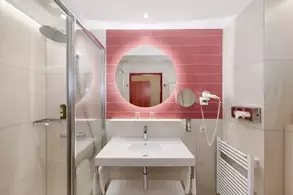 Bad mit modernen Elementen. Eine große Spiegelrunde ist über einem weißen Waschtisch platziert. Die Wände sind hell gefliest, mit pinkfarbener Akzentwand. Eine Dusche aus Glas ist sichtbar, und zusätzliche Badinstallationen sind vorhanden.