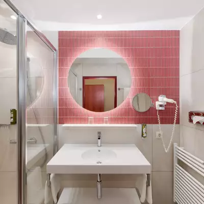 Bad mit modernen Elementen. Eine große Spiegelrunde ist über einem weißen Waschtisch platziert. Die Wände sind hell gefliest, mit pinkfarbener Akzentwand. Eine Dusche aus Glas ist sichtbar, und zusätzliche Badinstallationen sind vorhanden.